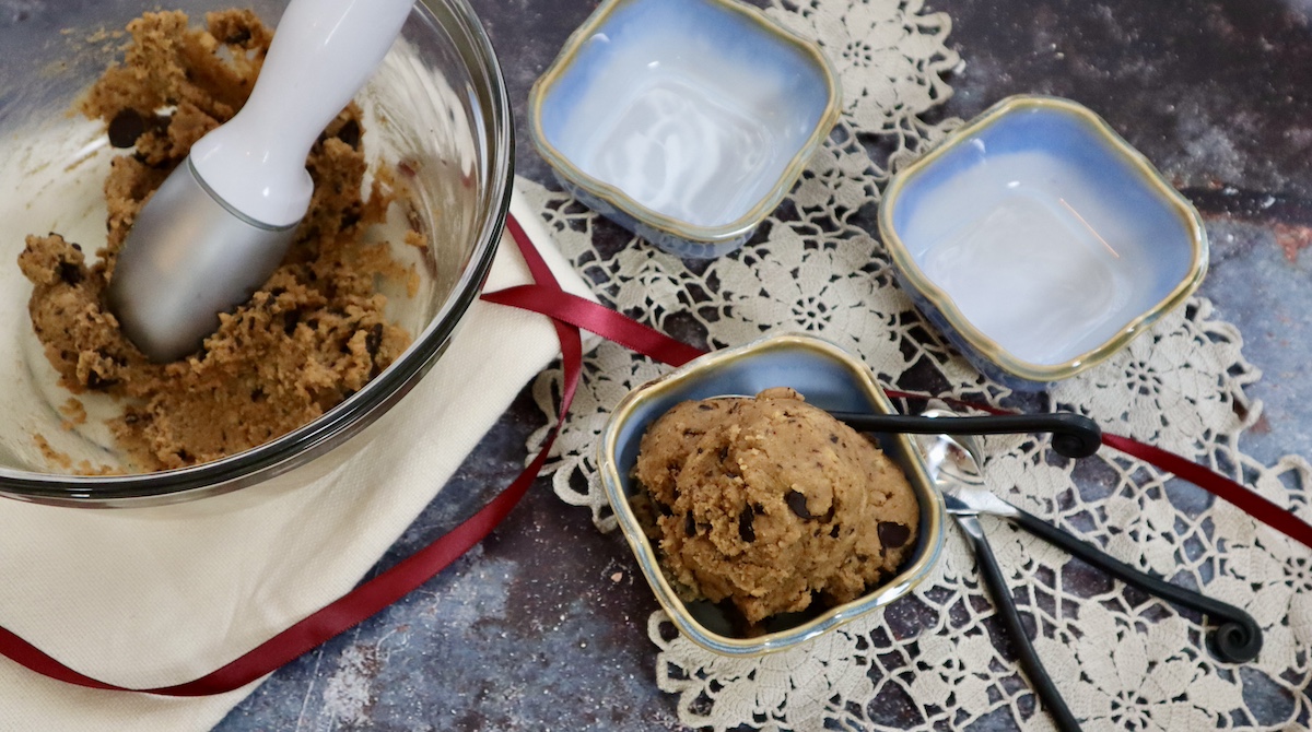 This is a photo of blue dessert dish filled with dairy and gluten free chickpea cookie dough with a large bowl filled with the dough and two more empty blue dishes with three black handled spoons on a lacy cloth.