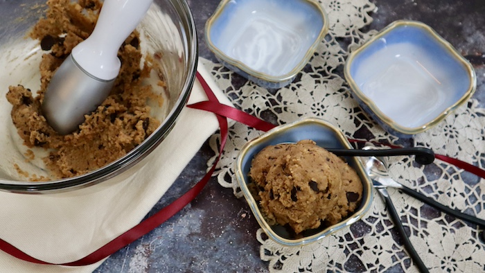 This is a photo of a square light blue dish filled with dairy and gluten free chickpea cookie dough with a big clear bowl filled with the cookie dough next to it and two other light blue square dishes and three black handled spoons on a lacy cloth.