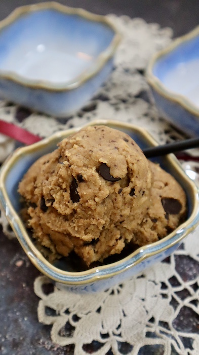 This is a photo of a small light blue square ceramic bowl with dairy and gluten free chickpea cookie dough in it with a little spoon with a black handle in it on a lacy cloth.
