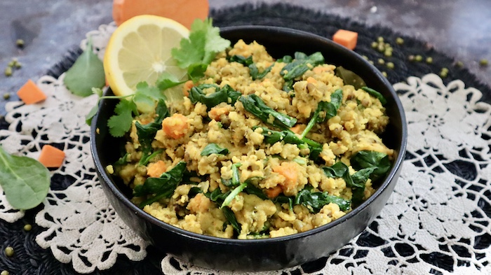 This is a photo of a black bowl filled with easy vegan green kitchari detox bowl on a white doily and black round table mat with sweet potato, fresh spinach and dried mung beans scattered near the bowl. 