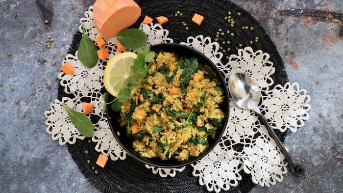 This is a photo of a black bowl filled with easy vegan green kitchari detox bowl on a white doily and black round table mat with sweet potato, fresh spinach and dried mung beans scattered near the bowl. 