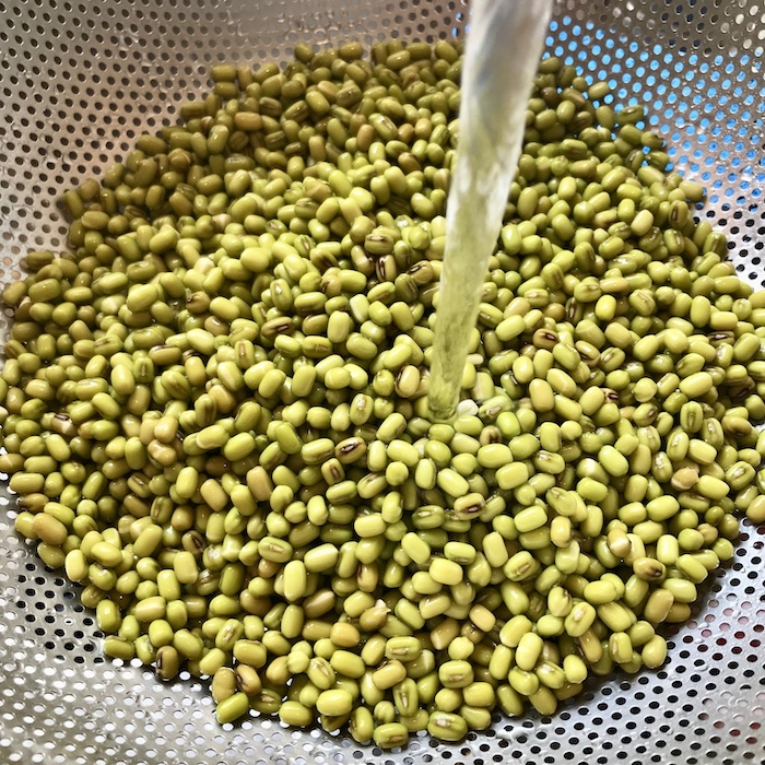 This is a photo of mung beans being rinsed with water in a metal sieve. 
