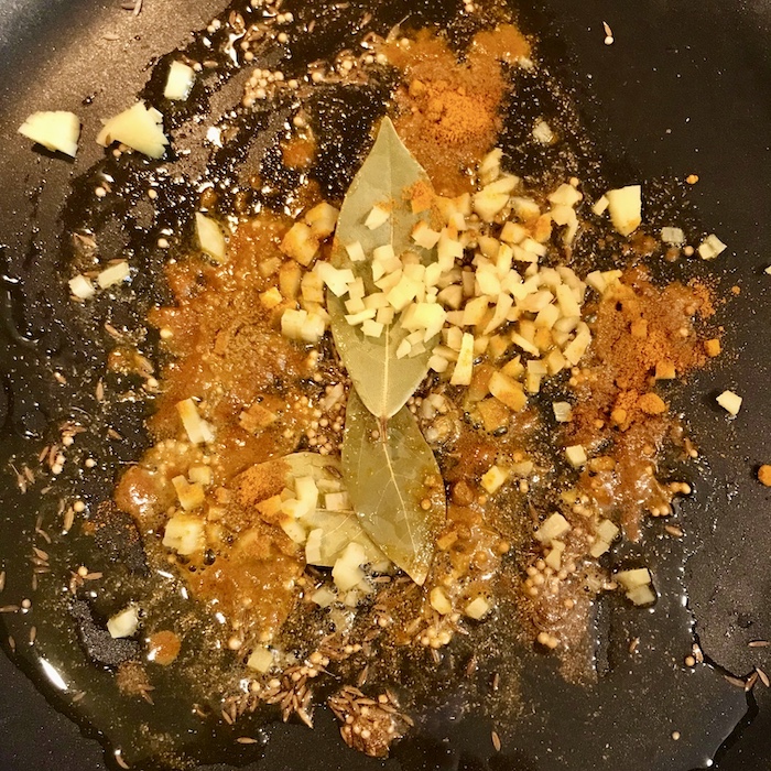 This is a photo of a skillet with oil and spices, minced garlic, chopped ginger and bay leaves in it.